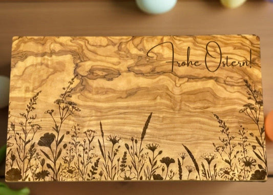 Ein rechteckiges Schneidebrett aus gemasertem Olivenholz liegt auf einem Holztisch. Auf der unteren Drittel des Bretts ist eine feine, schwarze Line-Art-Gravur mit verschiedenen Wiesenblumen und Gräsern zu sehen. Um das Brett herum liegen Tulpen in Frühlingsfarben, eine rote Tasse mit heller Flüssigkeit, ein pastellblaues Osterei und eine weiße Stoffserviette – alles in einer sanften, natürlichen Lichtstimmung.