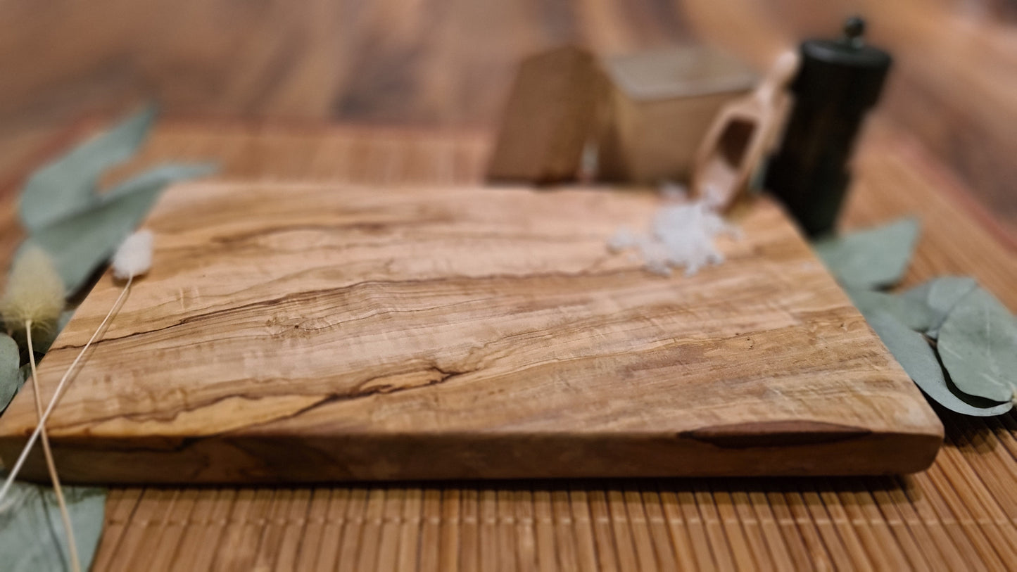 Ein rechteckiges Olivenholzbrett liegt auf einer Bambusunterlage. Auf dem Brett befinden sich grobes Meersalz und eine kleine Holzschaufel. Dahinter stehen ein Glasbehälter mit Salz, ein schwarzer Gewürzstreuer und ein Korkdeckel. Links und rechts vom Brett sind grüne Eukalyptusblätter sowie getrocknete Gräser dekorativ platziert. Die natürliche Maserung des Olivenholzes ist deutlich sichtbar und steht im Fokus des Bildes.