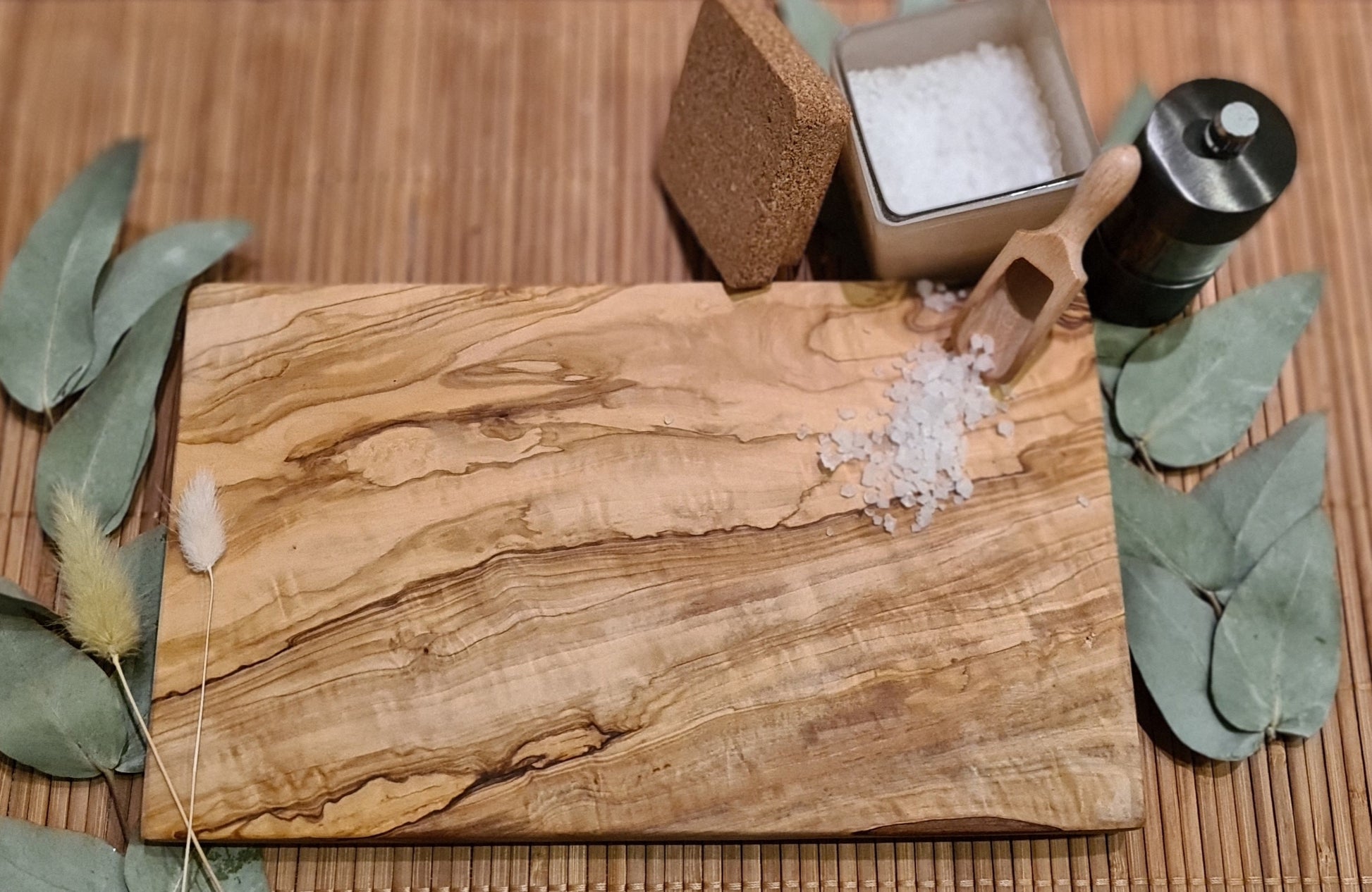 Ein rechteckiges Olivenholzbrett liegt auf einer Bambusunterlage. Auf dem Brett befinden sich grobes Meersalz und eine kleine Holzschaufel. Dahinter stehen ein Glasbehälter mit Salz, ein schwarzer Gewürzstreuer und ein Korkdeckel. Links und rechts vom Brett sind grüne Eukalyptusblätter sowie getrocknete Gräser dekorativ platziert. Die natürliche Maserung des Olivenholzes ist deutlich sichtbar und steht im Fokus des Bildes.