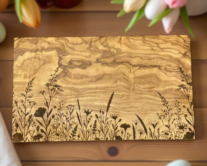Ein rechteckiges Schneidebrett aus gemasertem Olivenholz liegt auf einem Holztisch. Auf der unteren Drittel des Bretts ist eine feine, schwarze Line-Art-Gravur mit verschiedenen Wiesenblumen und Gräsern zu sehen. Um das Brett herum liegen Tulpen in Frühlingsfarben, eine rote Tasse mit heller Flüssigkeit, ein pastellblaues Osterei und eine weiße Stoffserviette – alles in einer sanften, natürlichen Lichtstimmung.